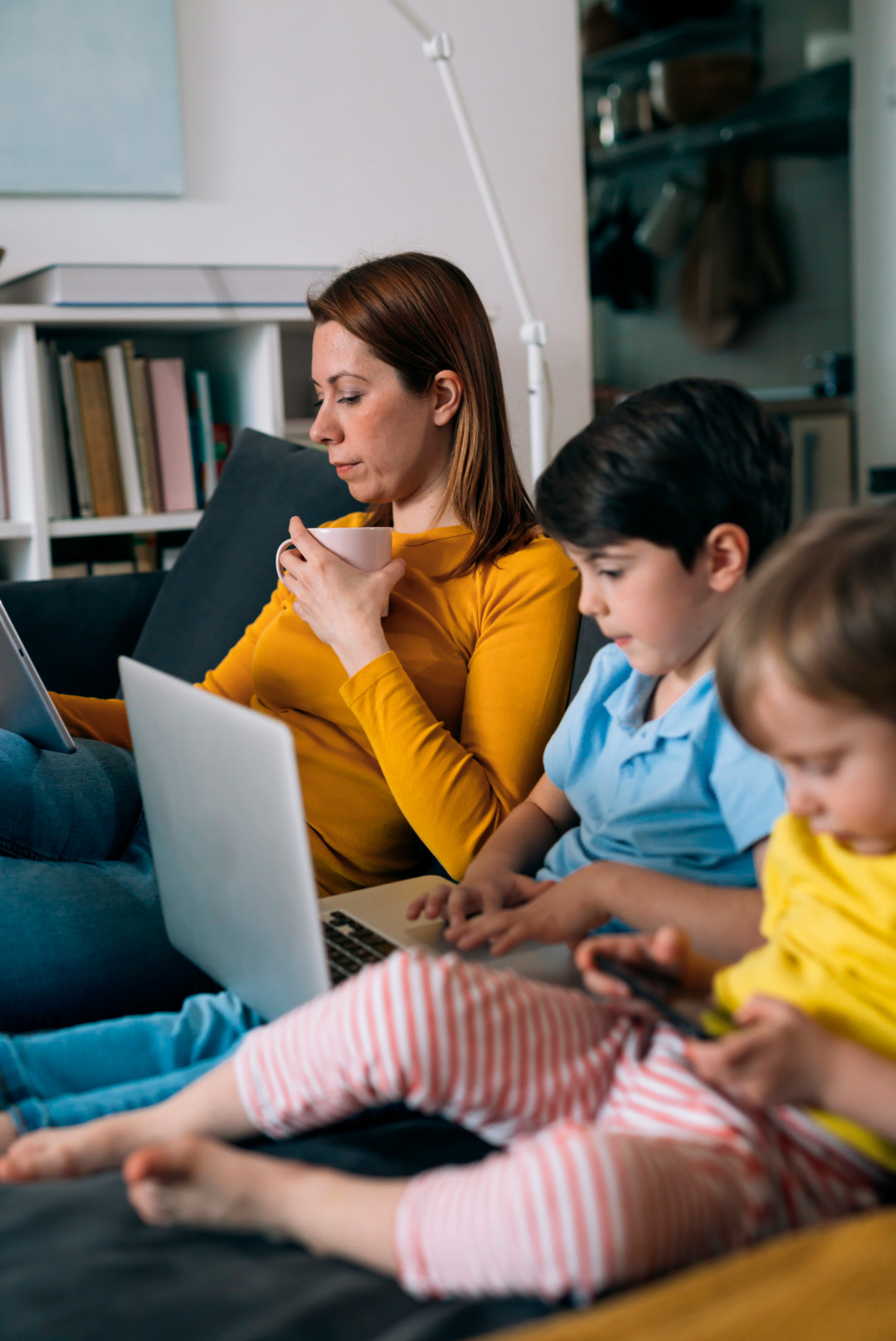 Madre con su laptop y un café, junto a sus hijos cada uno en su propio dispositivo